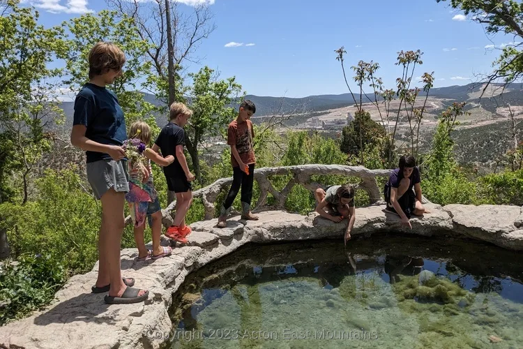 Students from Acton Academy East Mountains private school visiting Carlito Springs Open Space in Tijeras, New Mexico. They stand on the edge of a small pool of water with the Manzanito mountains in the background.